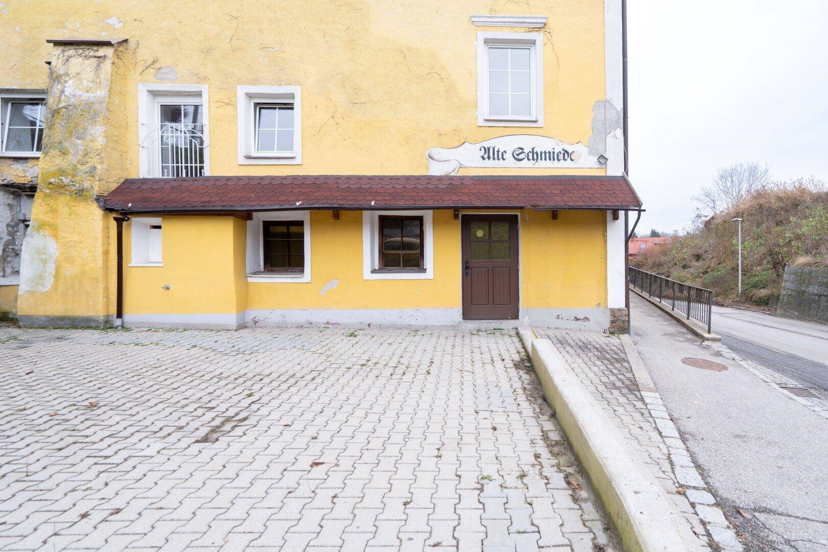 Gelbes historisches Gebäude mit gepflastertem Vorplatz und dem Schriftzug "Alte Schmiede".