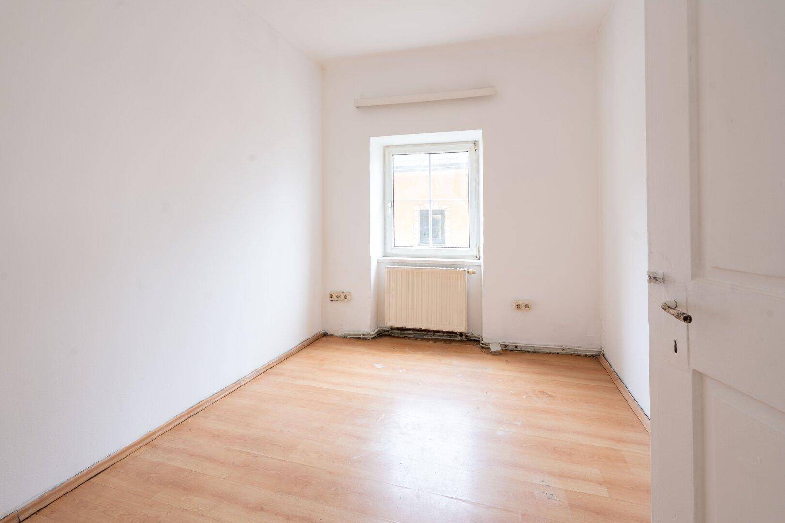 Leeres Zimmer mit hellem Laminatboden, weißen Wänden und einem Fenster mit Heizkörper darunter.