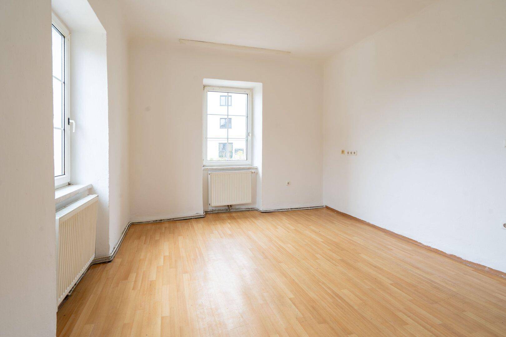 Geräumiges, helles Zimmer mit Laminatboden, weißen Wänden und zwei Fenstern.