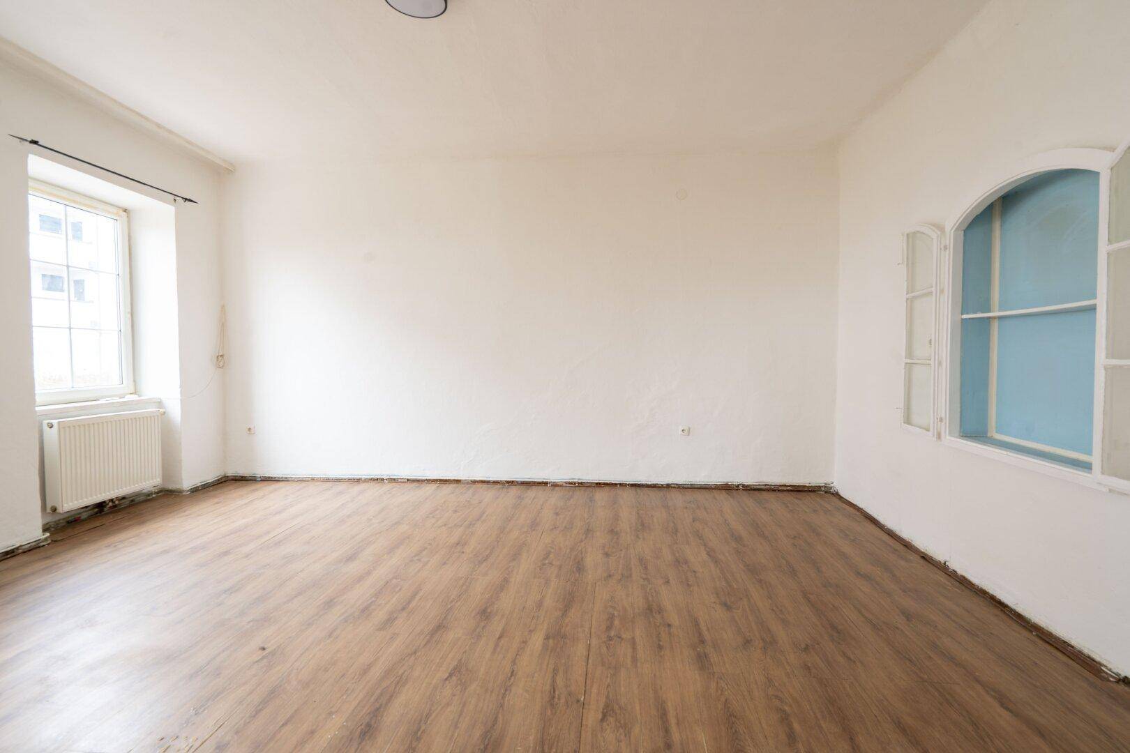 Helles Zimmer mit dunklem Laminatboden, weißen Wänden und einem dekorativen Bogenfenster.
