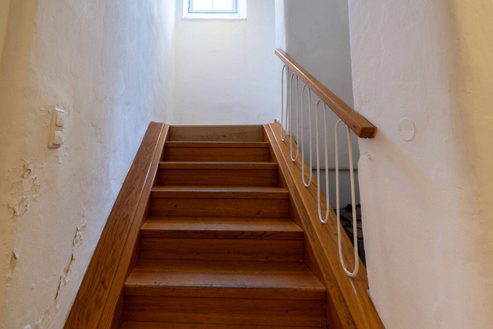 Holztreppe mit weißem Geländer führt in die obere Etage, Wände zeigen Abnutzungsspuren.