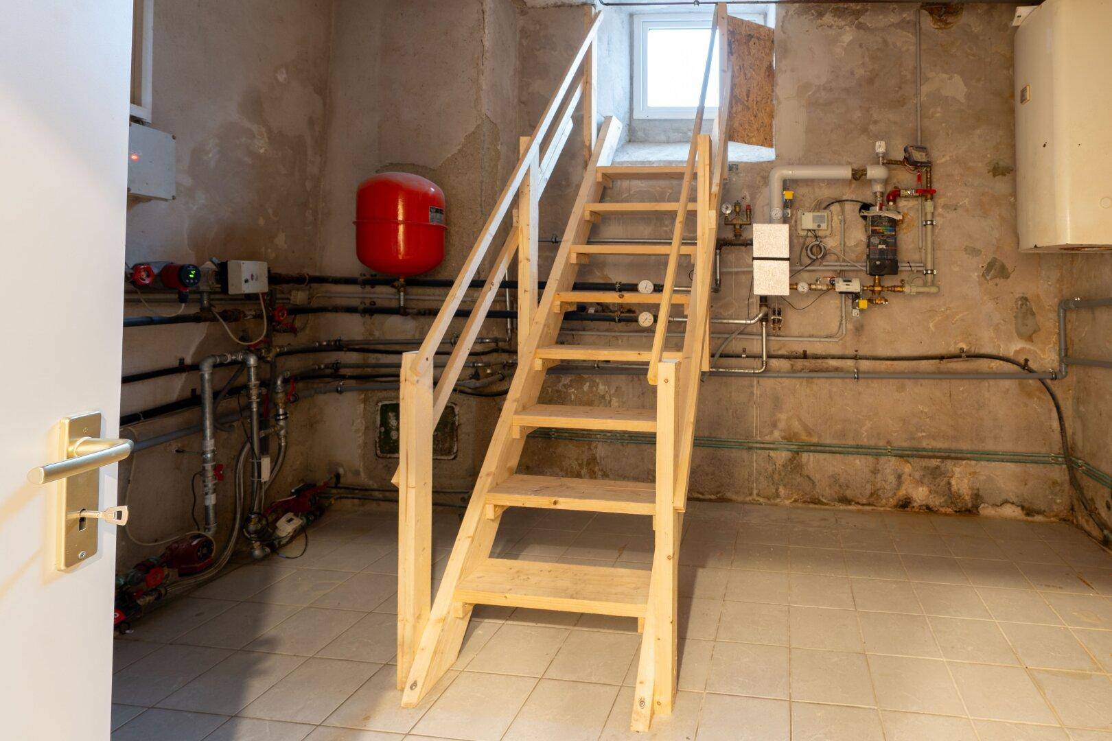 Kellerraum mit Heizungsanlage, sichtbaren Rohren und einer einfachen Holztreppe.