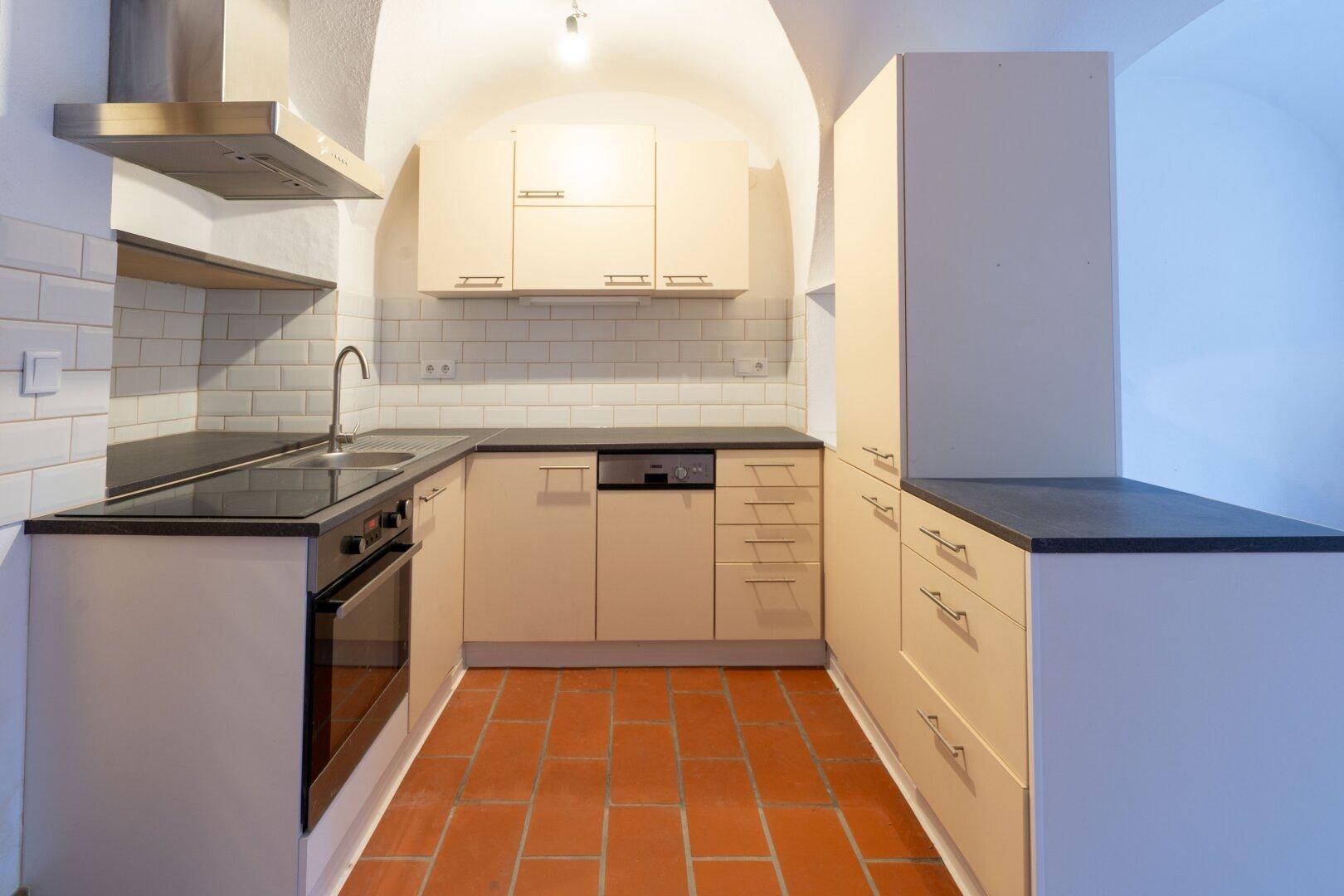 Moderne Küche mit beigen Schränken, dunkler Arbeitsplatte, Induktionskochfeld und Terrakottafliesen.