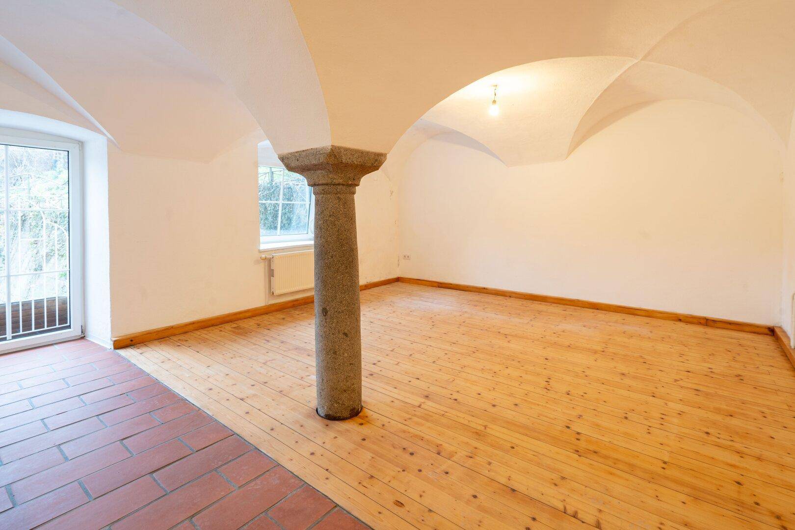Geräumiger Raum mit Gewölbedecke, Steinsäule, Holzdielenboden und Zugang nach draußen.