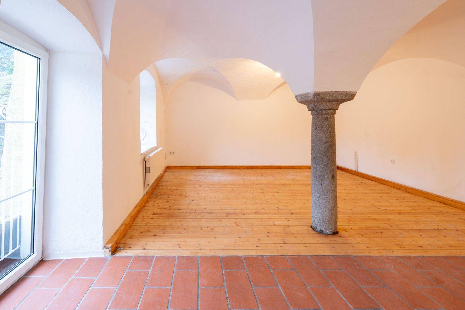Heller Raum mit Gewölbedecke, Steinsäule, Holzdielenboden und Zugang zu einem Außenbereich.