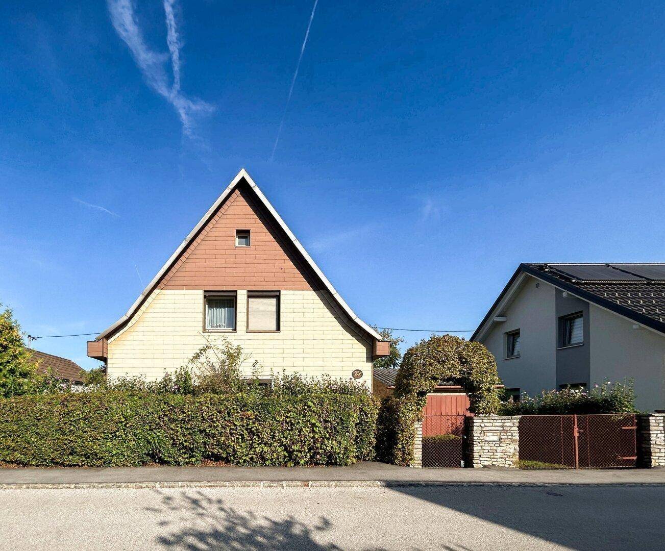 Hausfassade mit markantem Satteldach und gepflegter Hecke unter blauem Himmel.