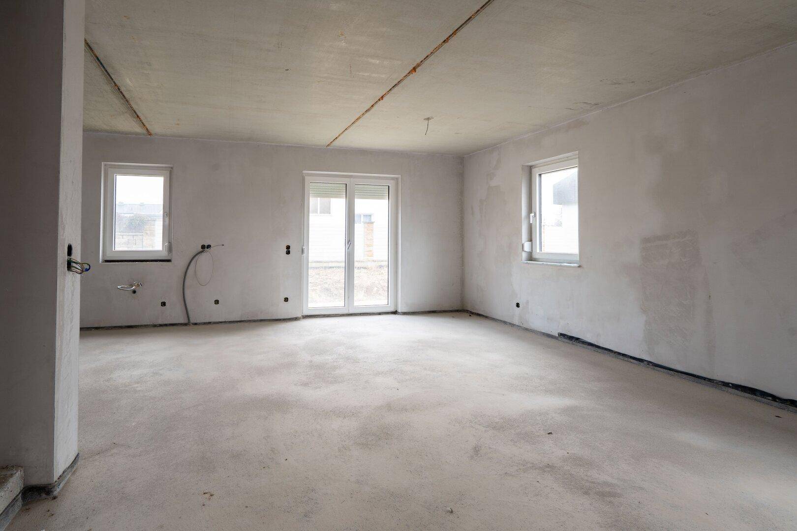 Geräumiger Wohnbereich im Rohbau mit Terrassentür, Fenstern und Betonboden.