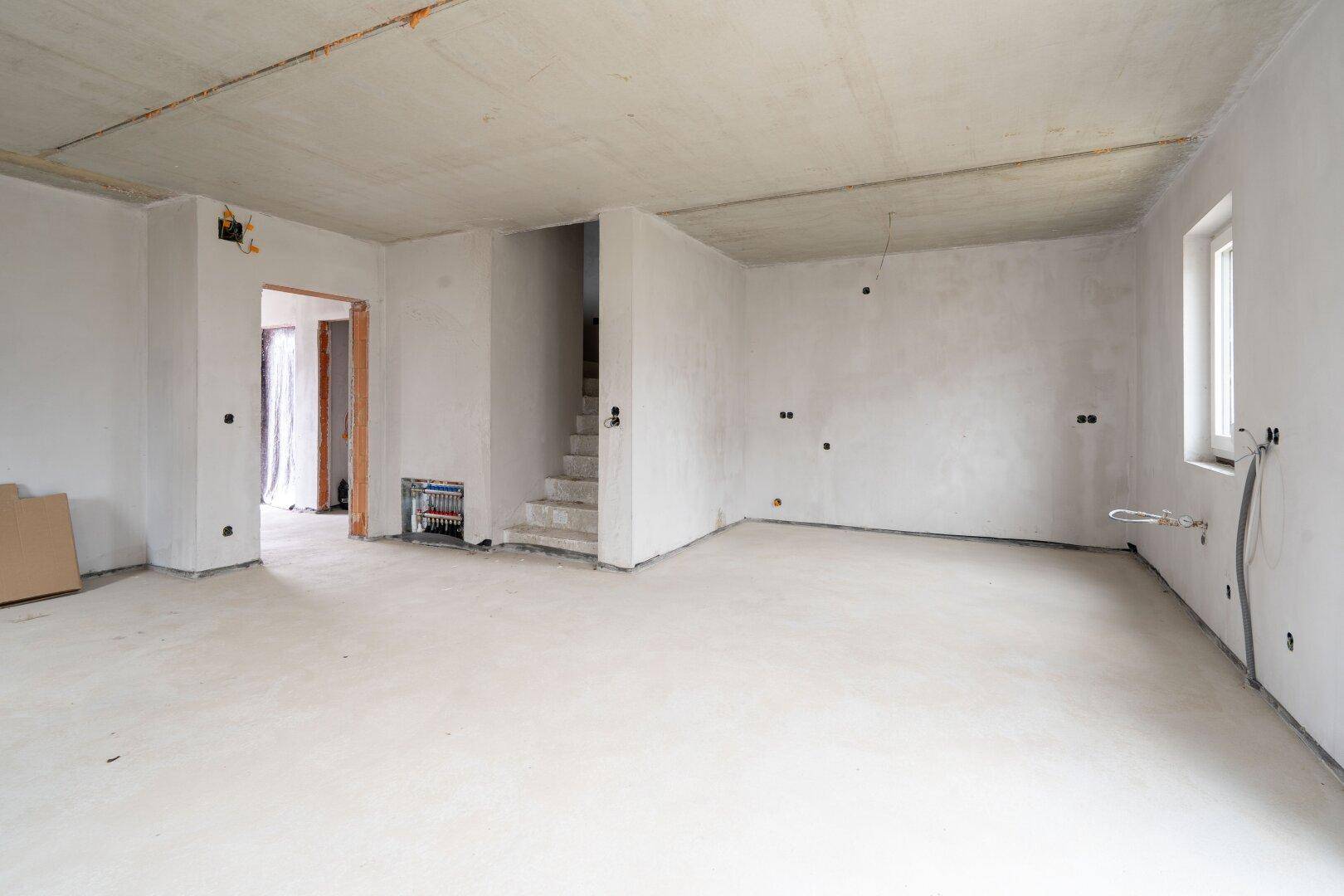 Großer Wohnbereich im Rohbau mit Treppenaufgang, Fenstern und Betonboden.