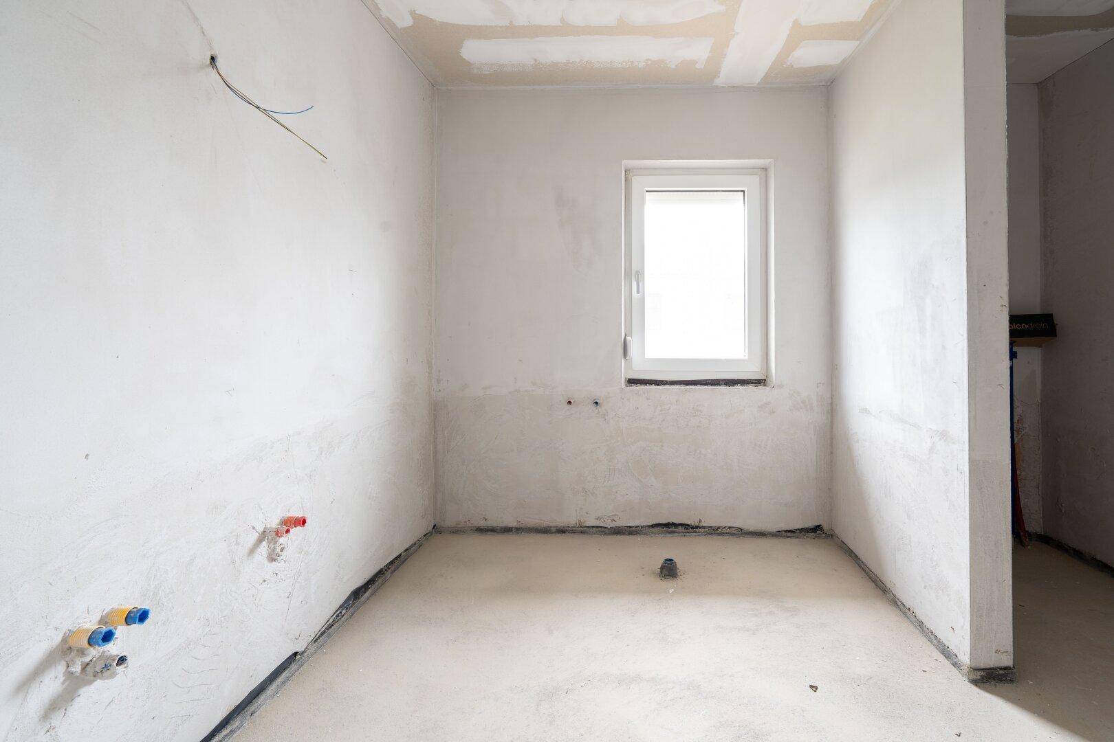 Badezimmer im Rohbau mit sichtbaren Wasseranschlüssen, Fenster und Betonboden.