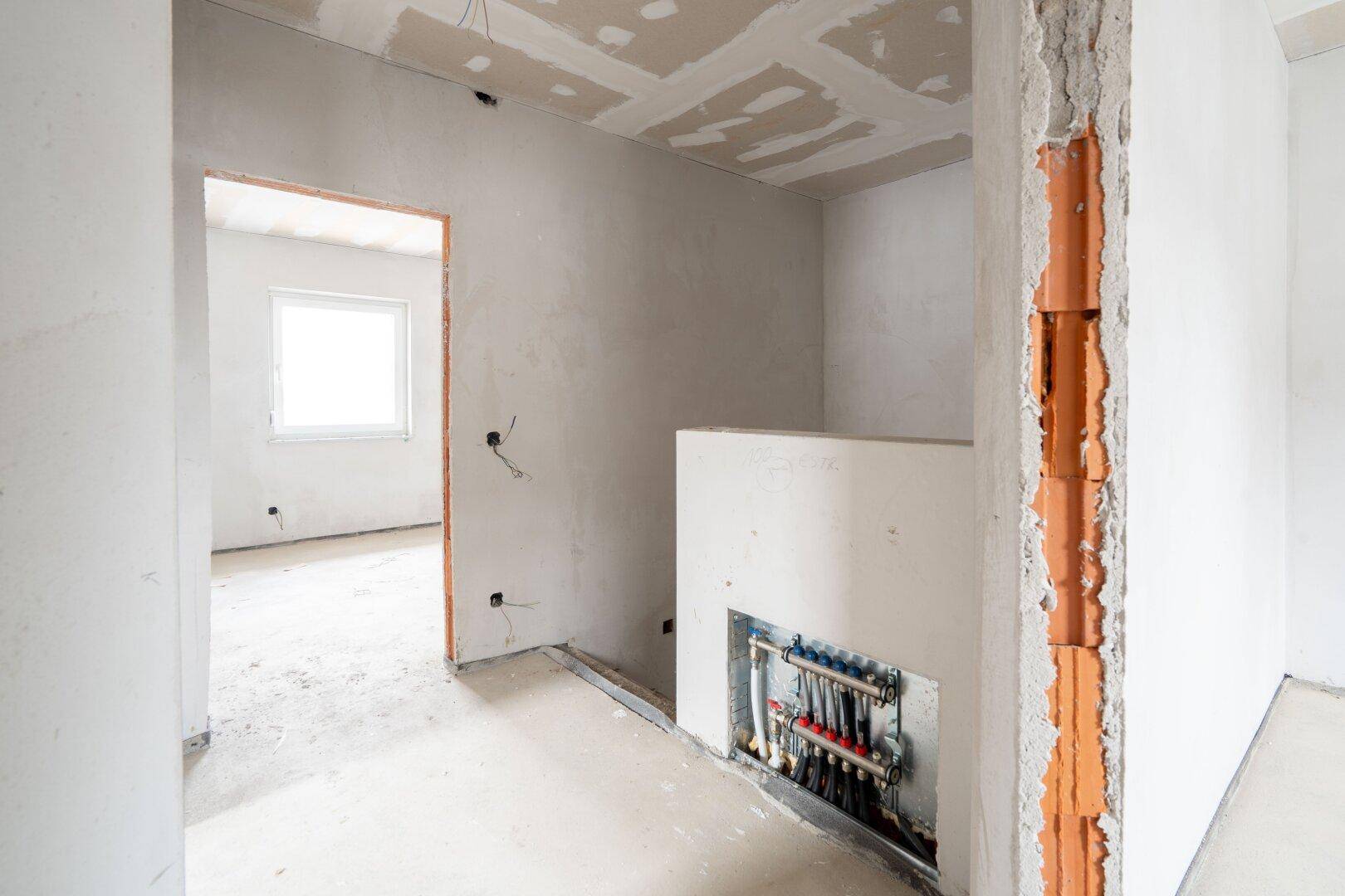 Flurbereich im Rohbau mit sichtbarem Heizverteiler und Übergang zu weiteren Räumen.
