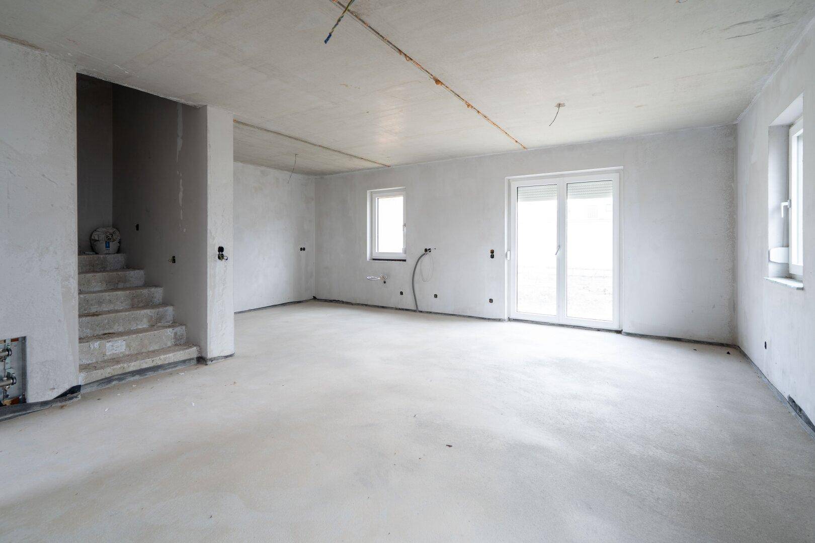 Geräumiger Wohnbereich im Rohbau mit Treppenaufgang, Terrassentür und Betonboden.