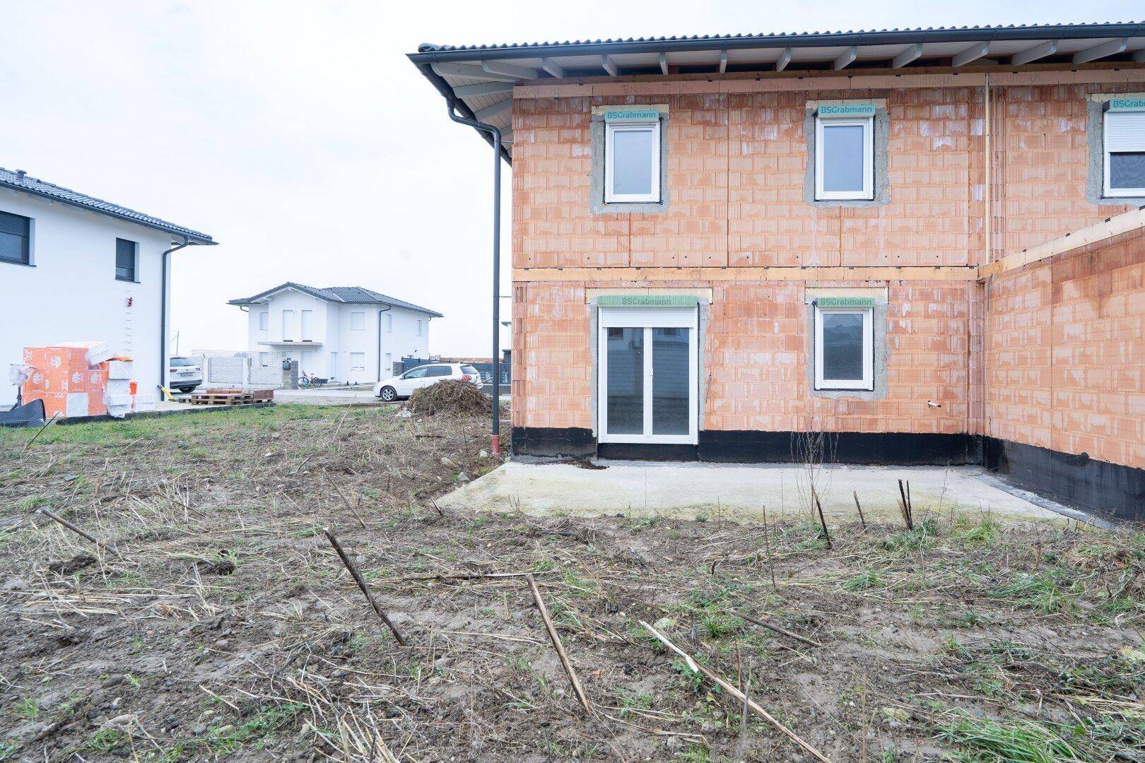Rückansicht der Doppelhaushälfte im Rohbau mit Terrassentür und ungestaltetem Außenbereich.