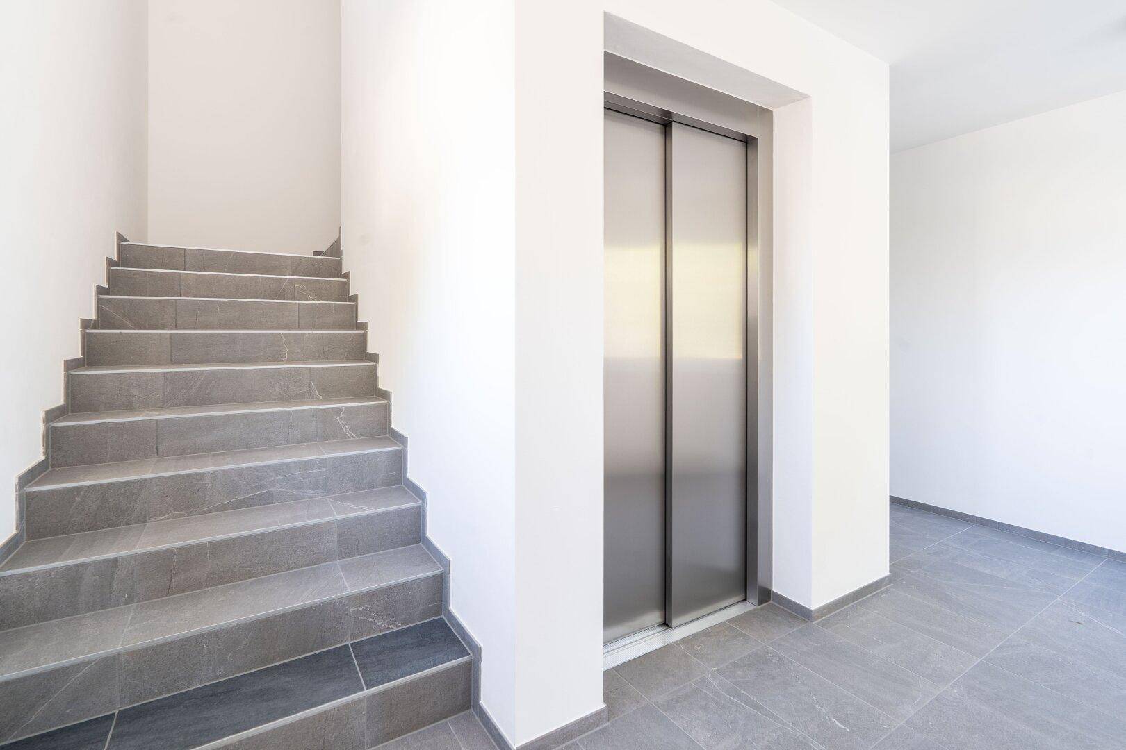 Treppenhaus mit grauen Fliesen und einem modernen Aufzug, der alle Etagen bequem und barrierefrei verbindet.