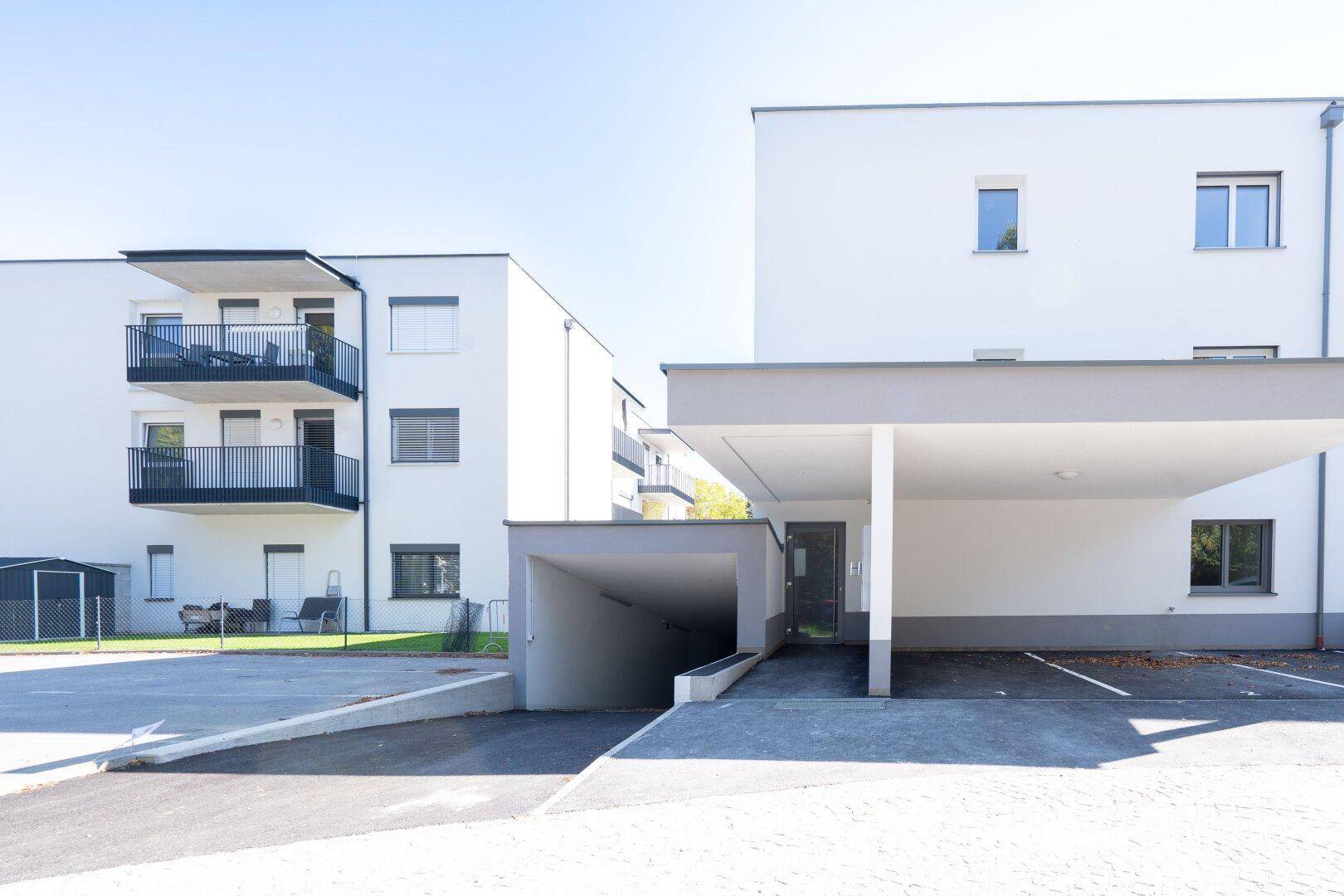 Außenansicht eines modernen Wohnkomplexes mit Tiefgaragenzufahrt und Parkplätzen im Freien.