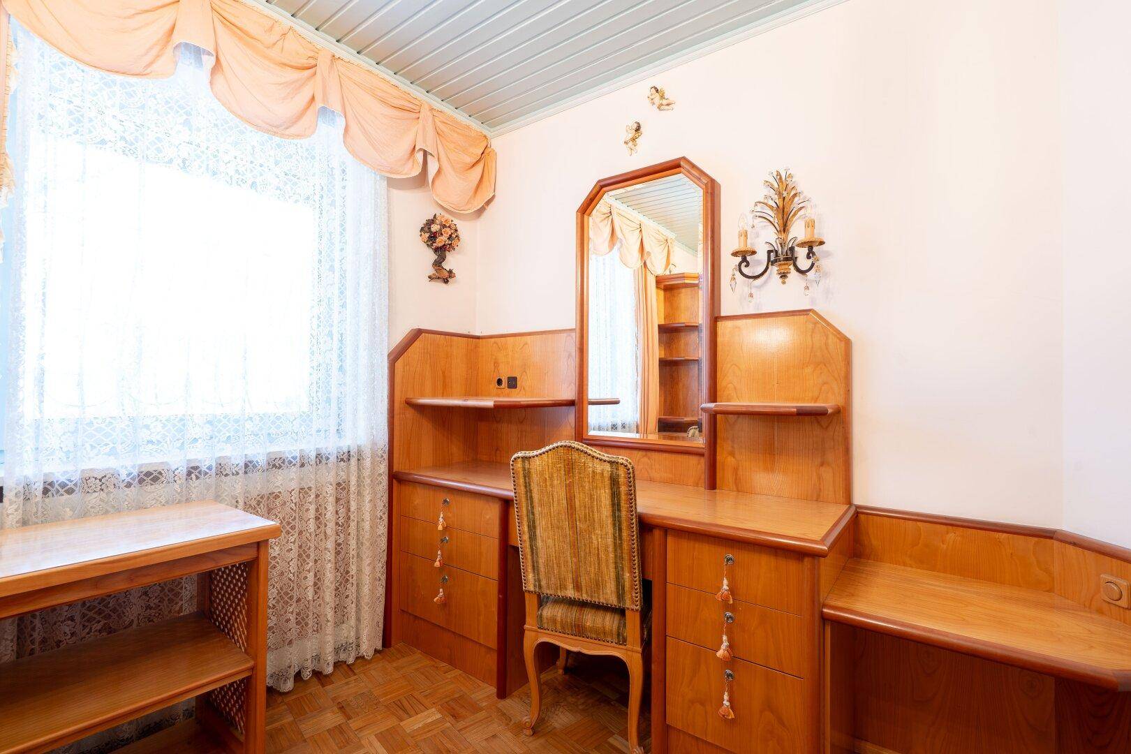 Schlafzimmer mit Holzmöbeln, Schminktisch und großem Fenster, das viel Tageslicht hereinlässt.