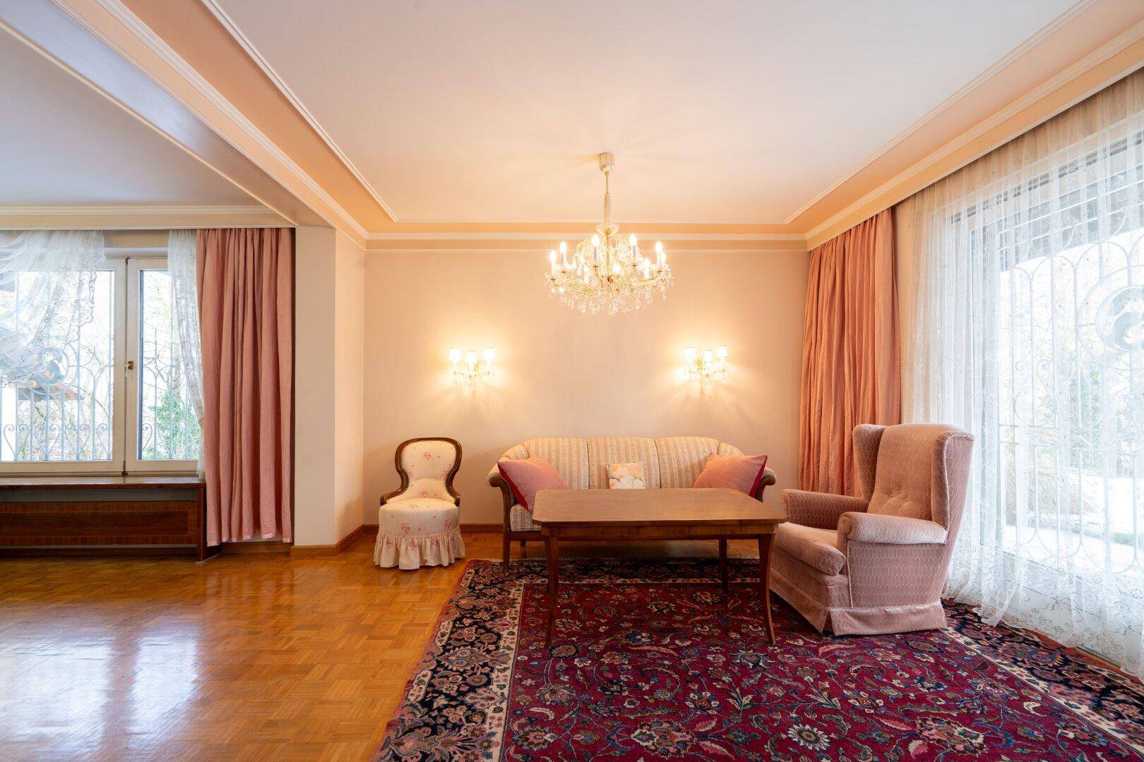 Geräumiges Wohnzimmer mit Parkettboden, Kronleuchter und großen Fenstern, die den Raum erhellen.
