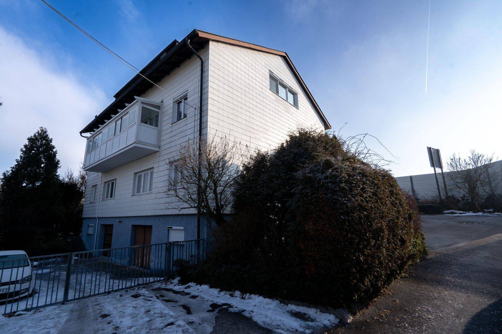 Seitenansicht des Wohngebäudes mit weißer Fassade und verglaster Loggia, umgeben von Bäumen und einem Zaun.