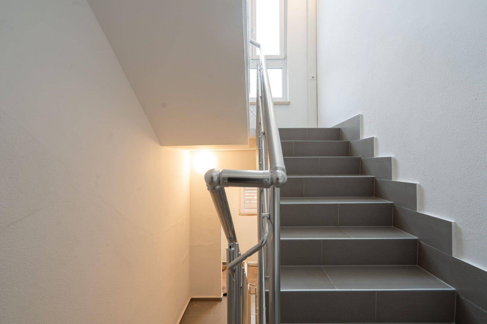 Modernes Treppenhaus mit grauen Fliesenstufen und einem eleganten Edelstahlgeländer in hellem Ambiente.