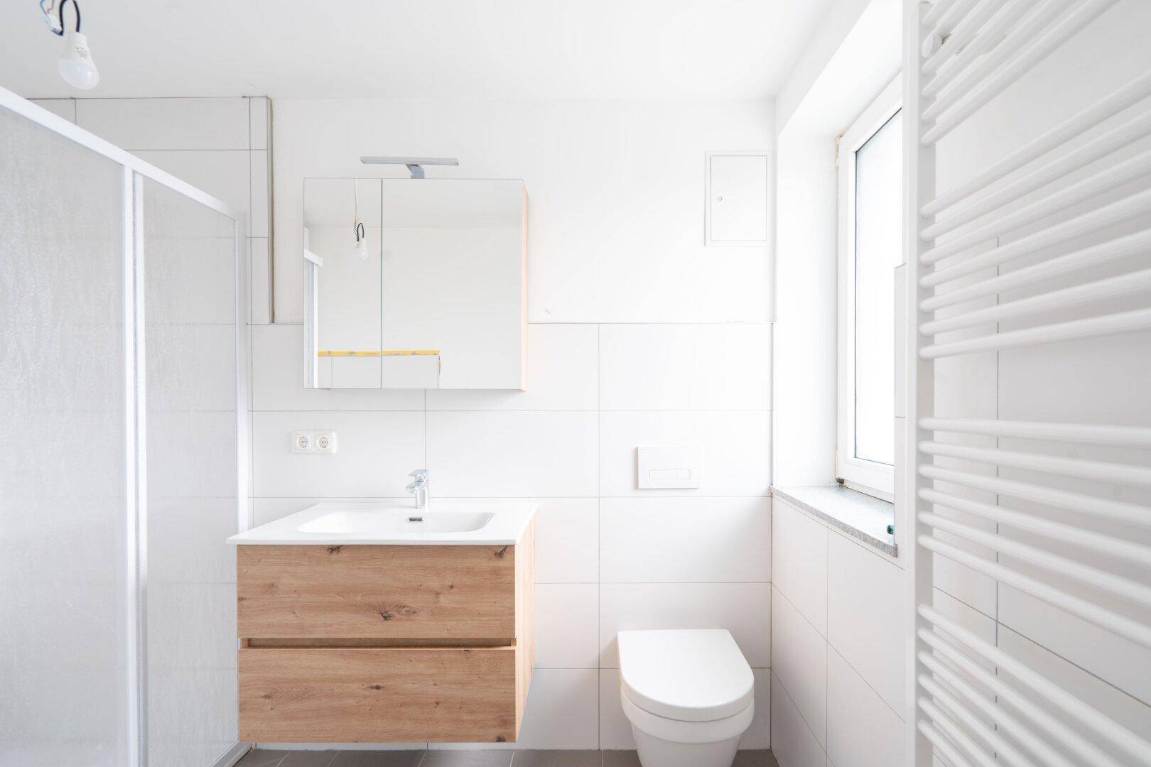 Modernes Badezimmer mit weißen Fliesen, Holzwaschtisch, Duschkabine und einem Fenster für Tageslicht.