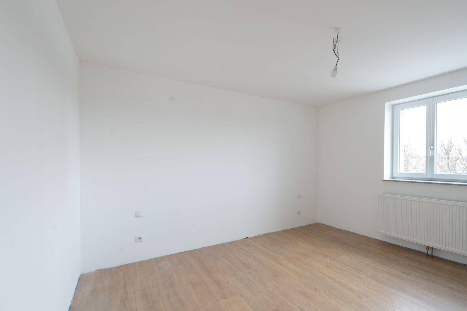 Leeres Schlafzimmer mit hellem Laminatboden und einem Fenster, das viel Tageslicht hereinlässt.