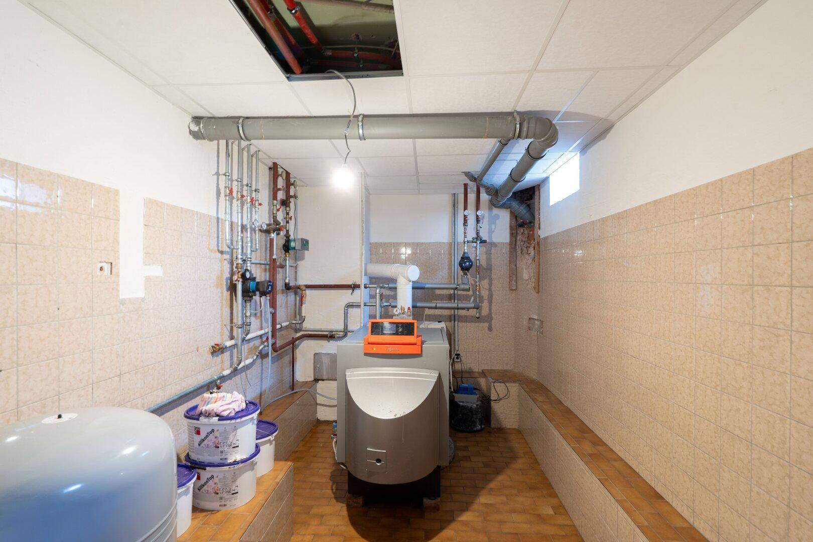 Heizungsraum mit umfangreicher Installation von Rohren und einem großen Heizkessel an gefliesten Wänden.