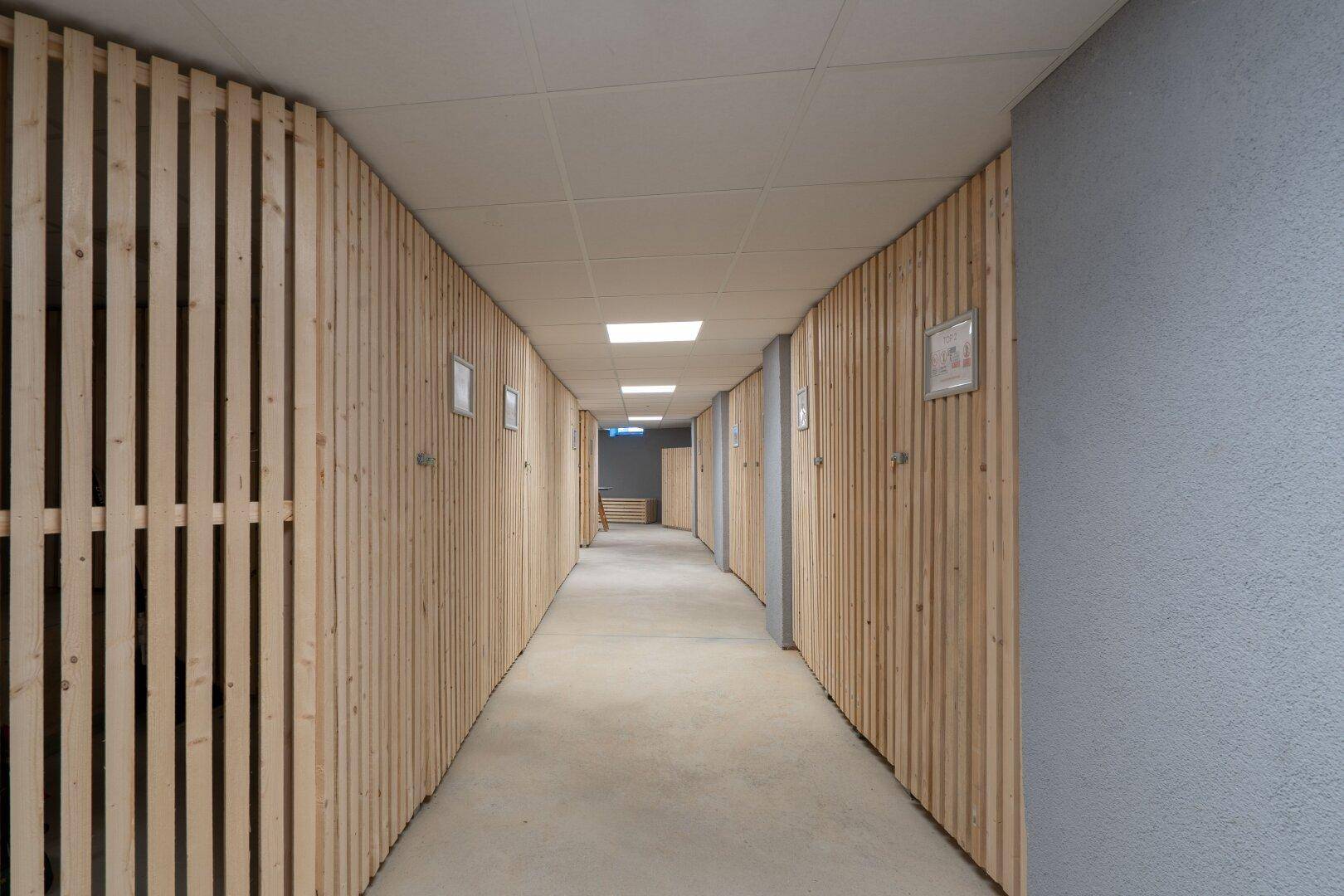 Langer Kellerflur mit Holzlattenwänden und Betonboden, der zu individuellen Lagerräumen führt.