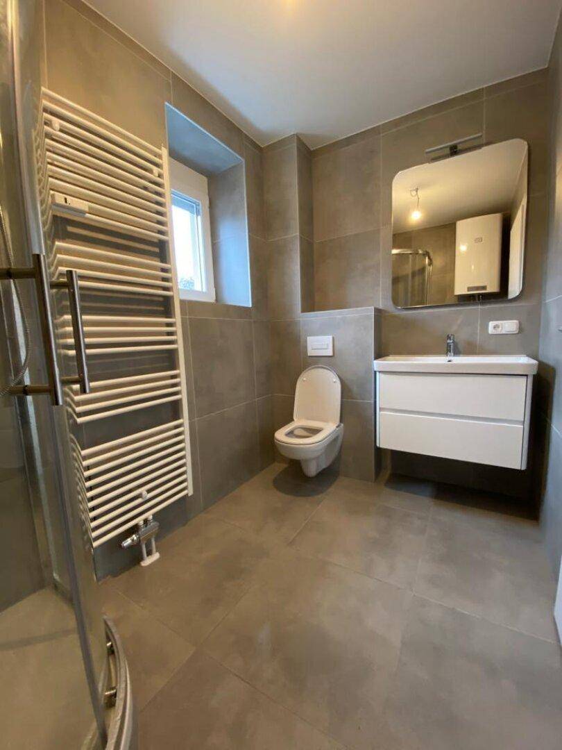 Modernes Badezimmer mit grauen Fliesen, Duschkabine und beheiztem Handtuchhalter sowie Fenster.