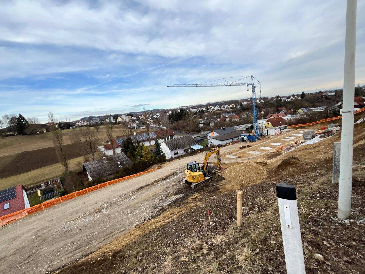 Baustelle mit Bagger und Kran, umgeben von einer Wohnsiedlung und ländlicher Landschaft unter bewölktem Himmel.