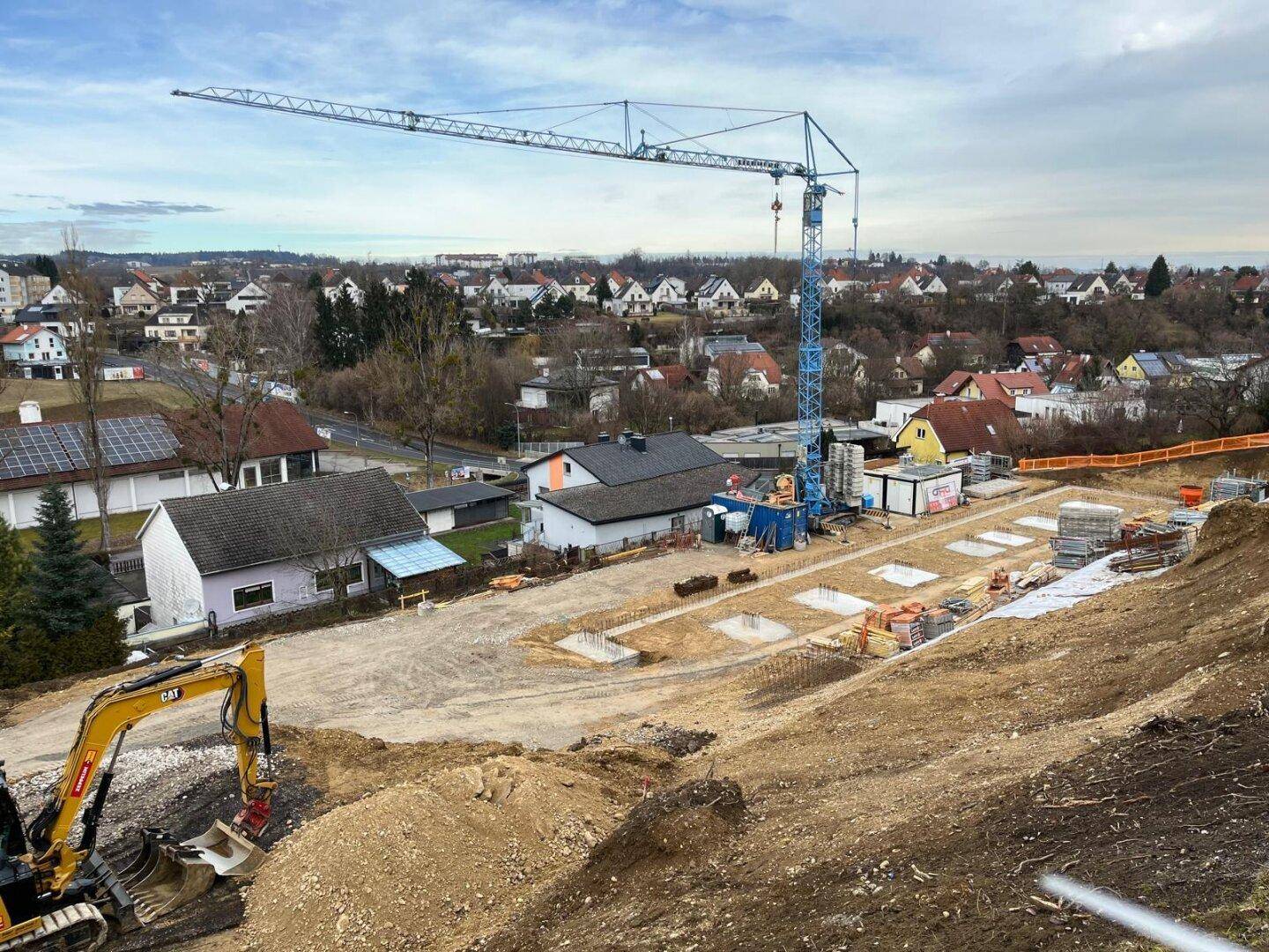 Übersicht der Baustelle mit sichtbaren Fundamenten, einem großen Kran und angrenzenden Wohnhäusern in der Ferne.