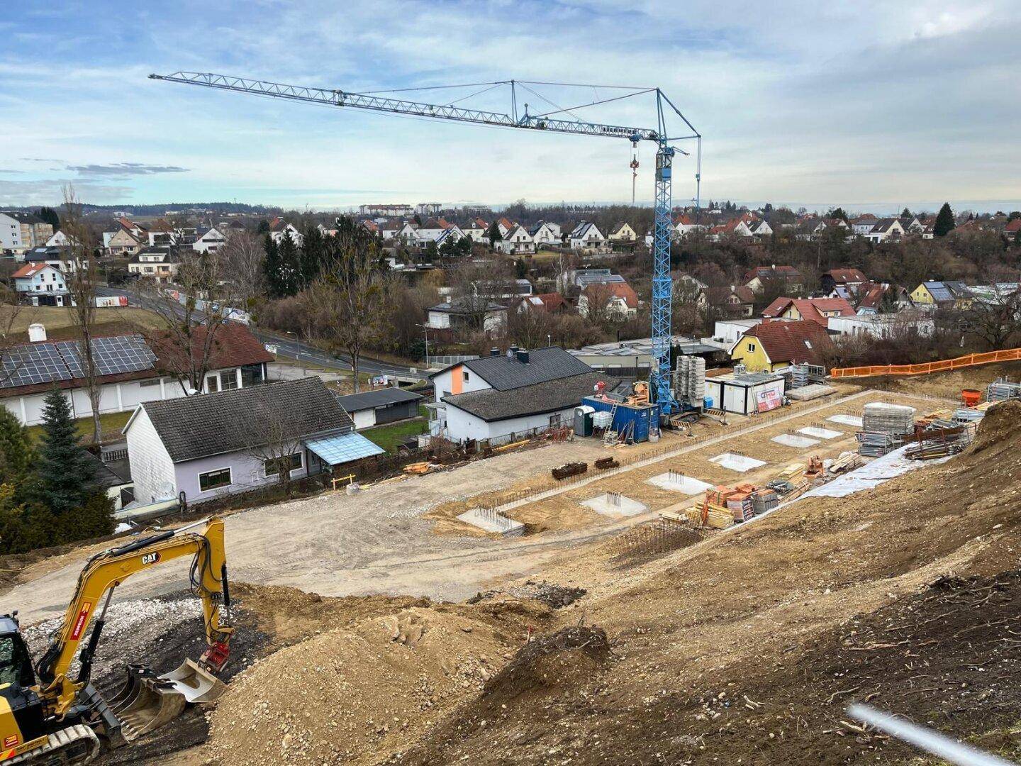 Detailansicht der Baustelle mit ausgehobenen Fundamenten, einem hohen Kran und umliegenden Häusern mit Solaranlagen.