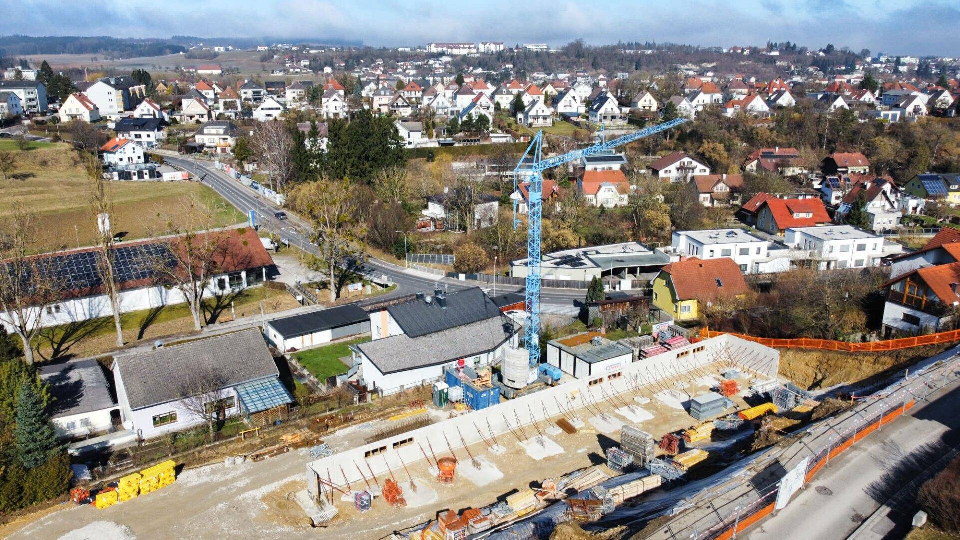 Luftaufnahme einer Baustelle in einem Wohngebiet mit Kran und umliegenden Häusern unter blauem Himmel.