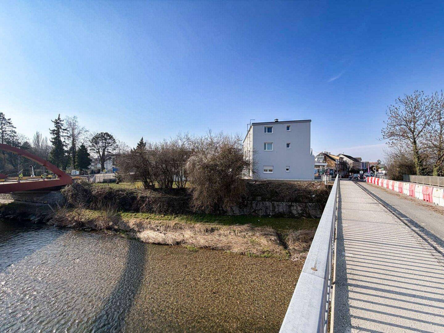 Gebäudeansicht von einer Brücke über einen Fluss, umgeben von Bäumen und Natur unter blauem Himmel.