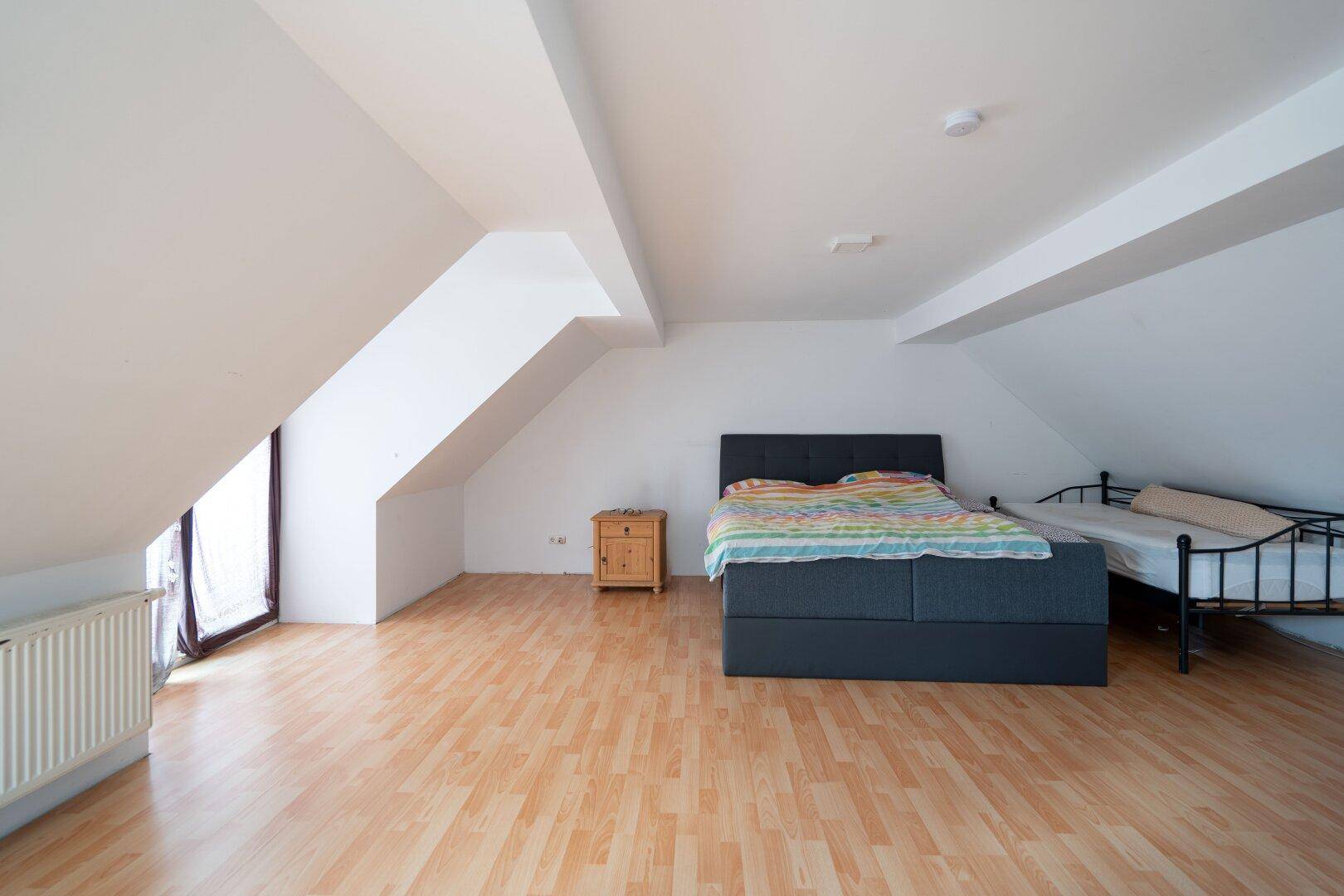 Helles Schlafzimmer im Dachgeschoss mit schrägen Wänden, Holzboden und großem Fenster für viel Licht.