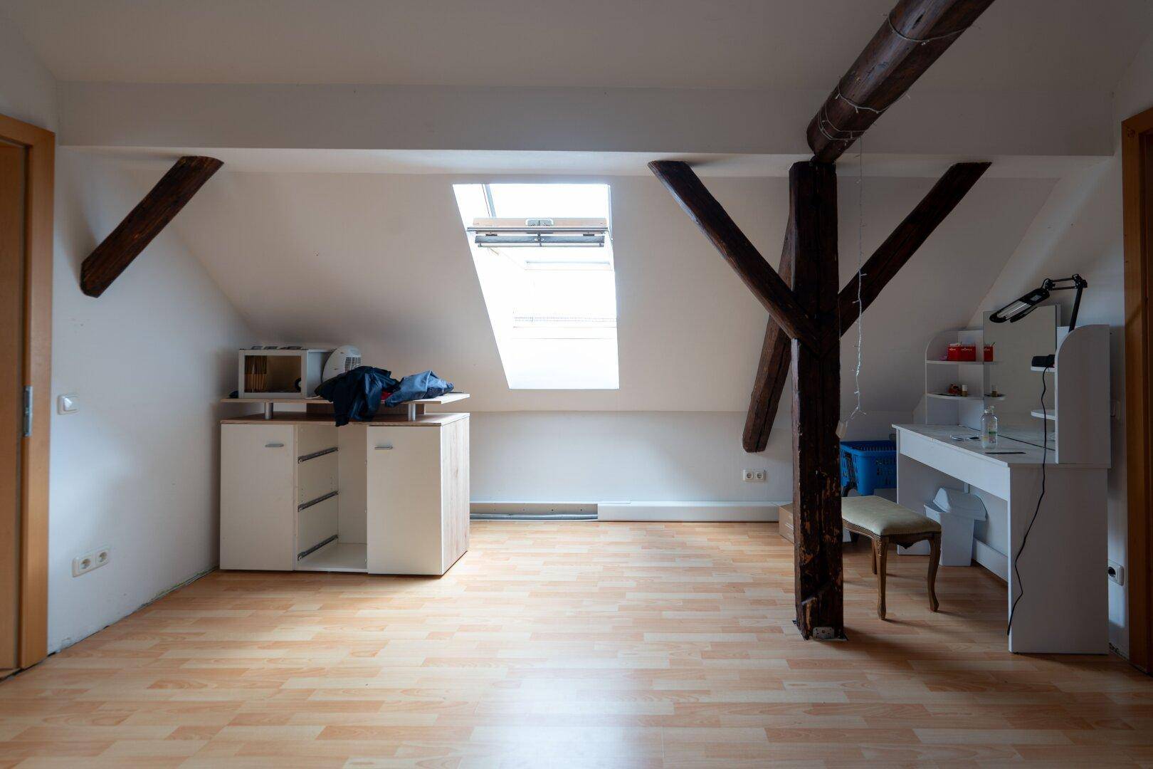 Dachgeschossraum mit Holzbalken, Dachfenster und einfacher Einrichtung als Arbeitsbereich oder Hobbyraum.