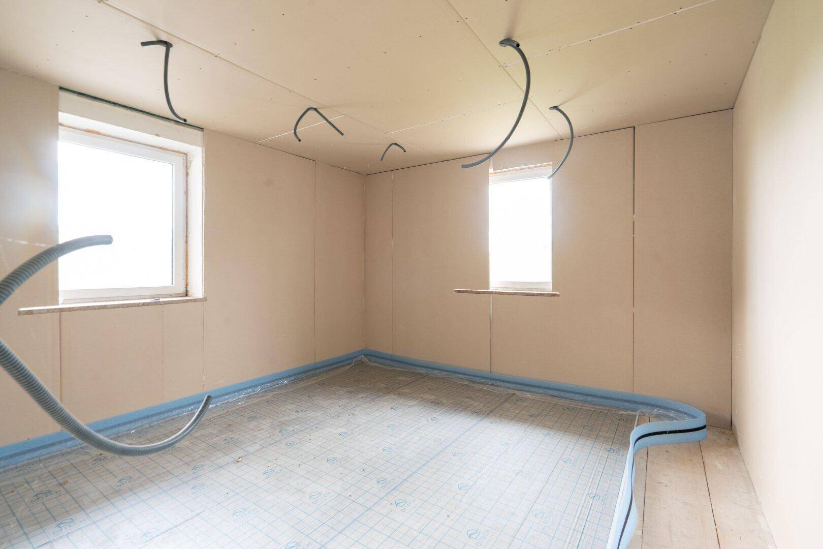 Raum im Rohbau mit vorbereitetem Boden für Fußbodenheizung und sichtbaren Leitungen für die Installation.