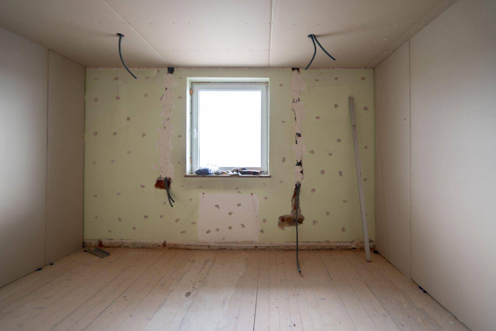 Raum im Rohbau mit Fenster, vorbereiteten Wänden und sichtbaren Elektroleitungen für zukünftige Nutzung.