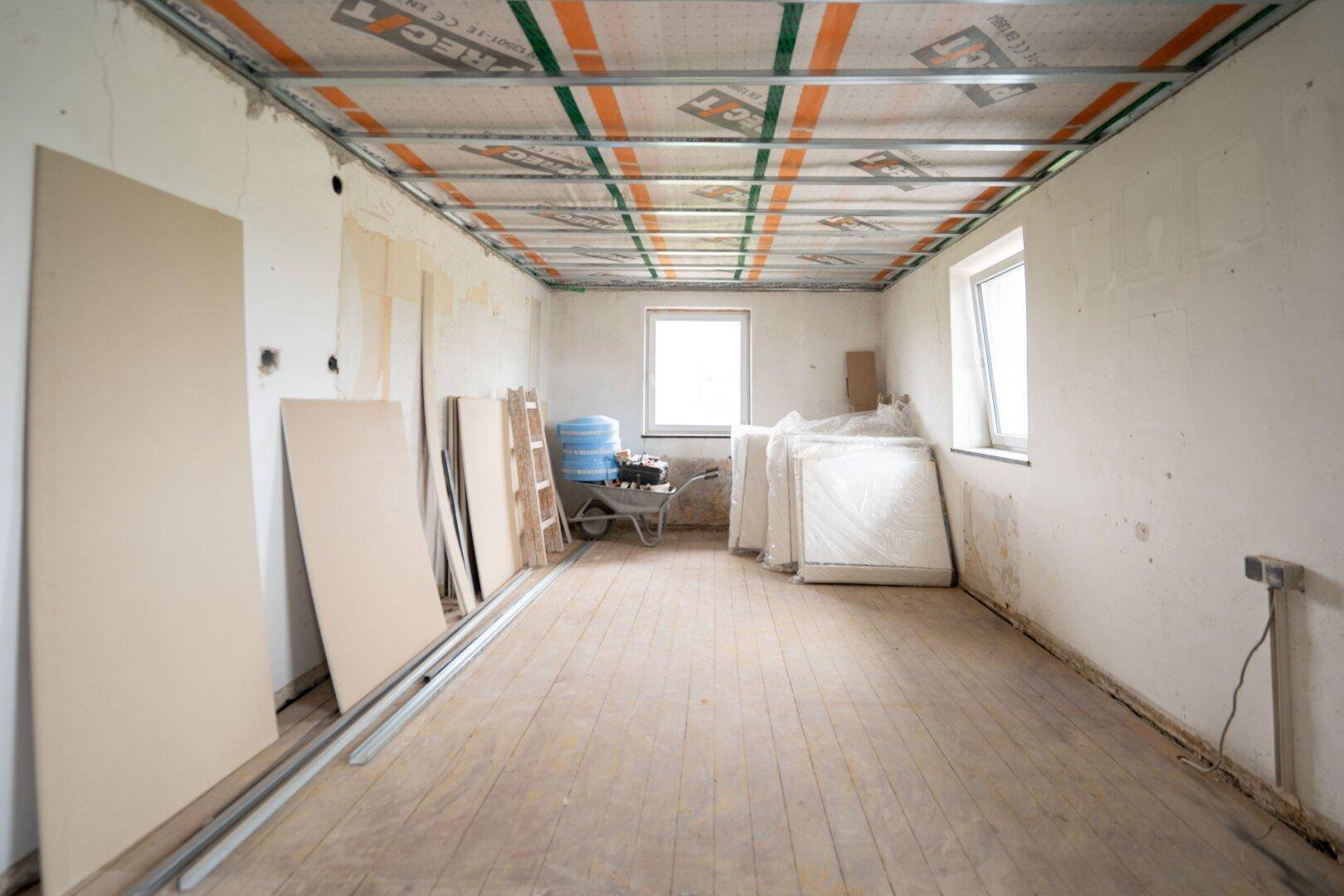Raum im Rohbau mit sichtbarer Deckenkonstruktion, Fenstern und Baumaterialien, bereit für den Ausbau.