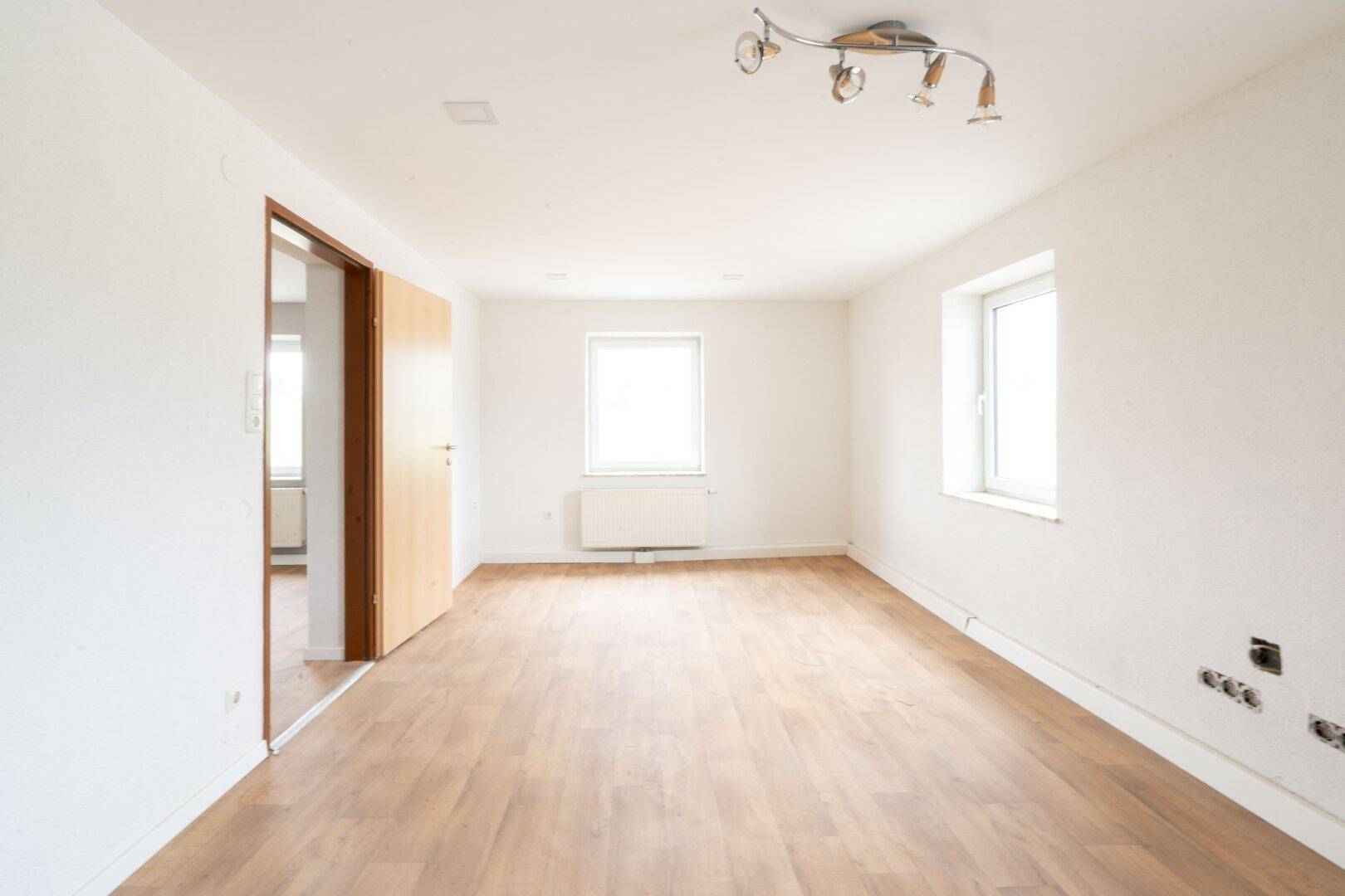 Heller, leerer Raum mit Holzboden, weißen Wänden und zwei Fenstern, die für viel Tageslicht sorgen.