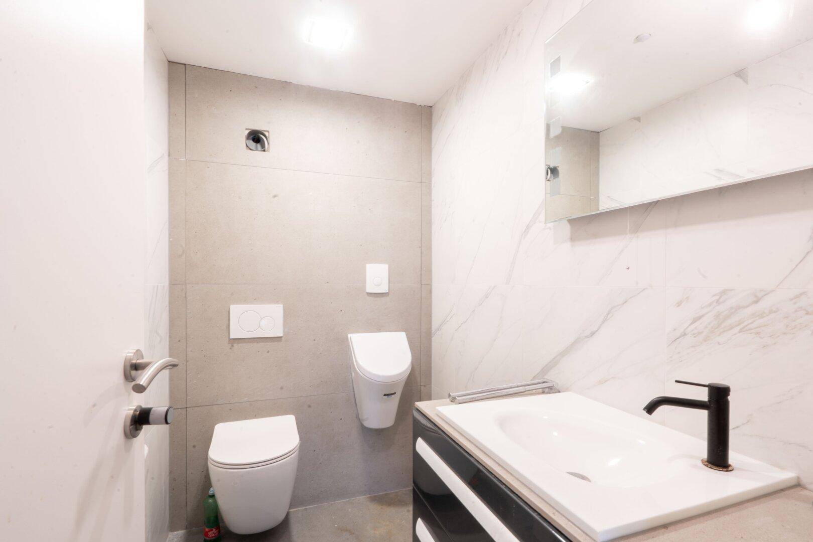 Modernes WC mit Wand-WC, Urinal, Waschbecken und eleganten Marmorfliesen an den Wänden.