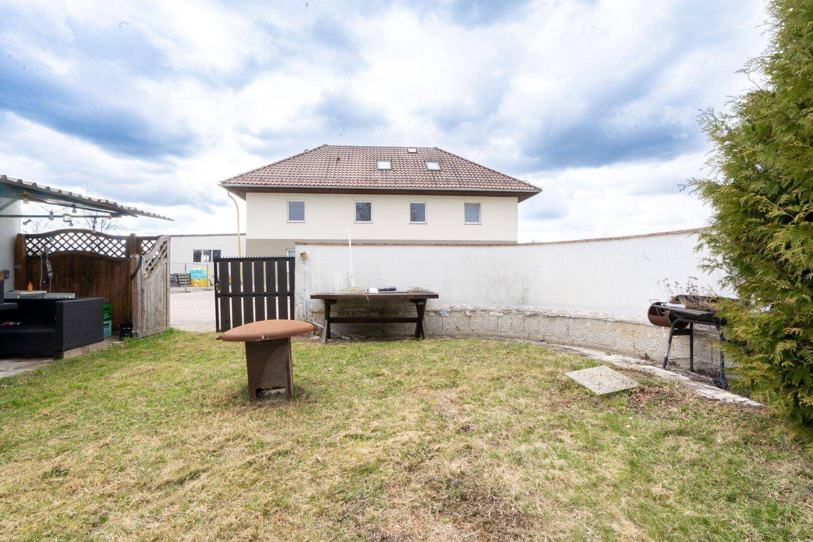 Garten mit Rasenfläche, Grillplatz und Holztisch, umgeben von einer niedrigen Mauer.