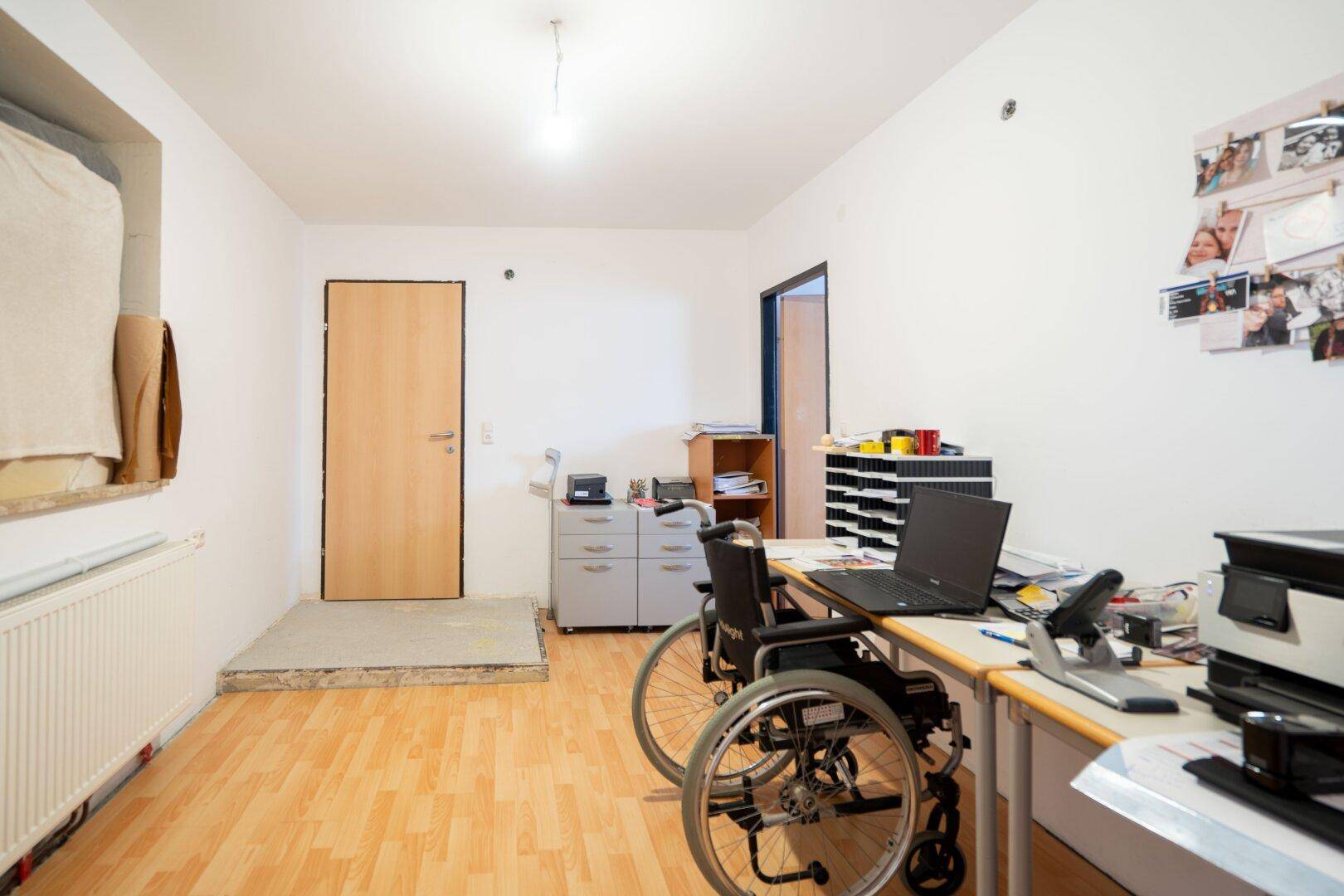 Helles Büro mit Holzboden, Heizkörper und einem Rollstuhl am Schreibtisch, funktional eingerichtet.