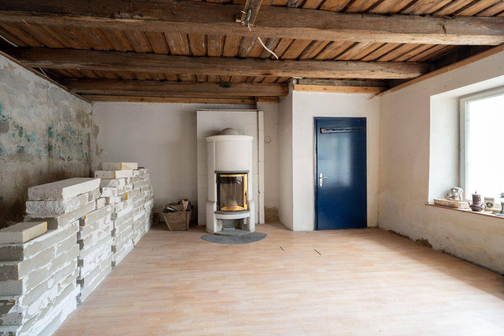 Renovierungsbedürftiger Raum mit freiliegenden Holzbalken und einem Kachelofen.