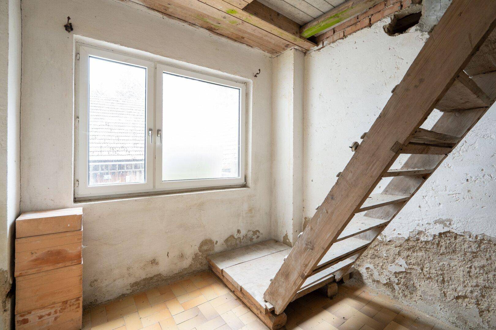 Unfertiger Raum mit einfacher Holztreppe und Fenster, bereit für den Ausbau.