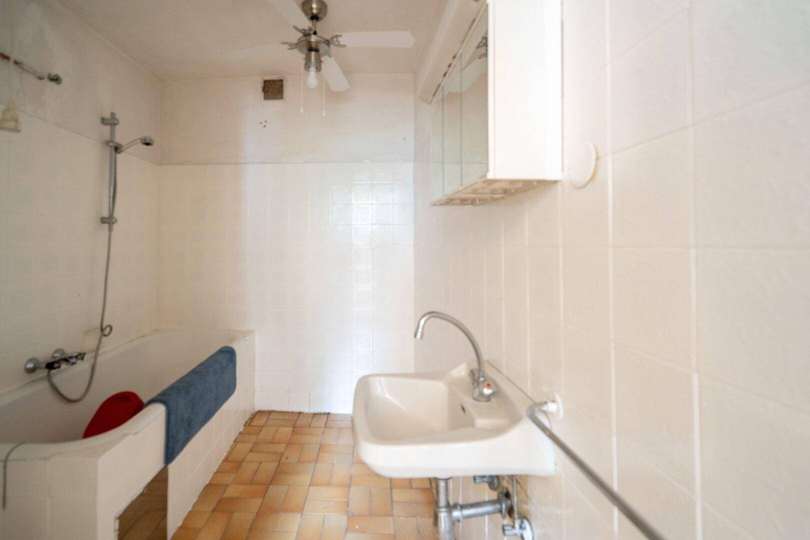 Einfaches Badezimmer mit Badewanne, Waschbecken und Deckenventilator, geflieste Wände.