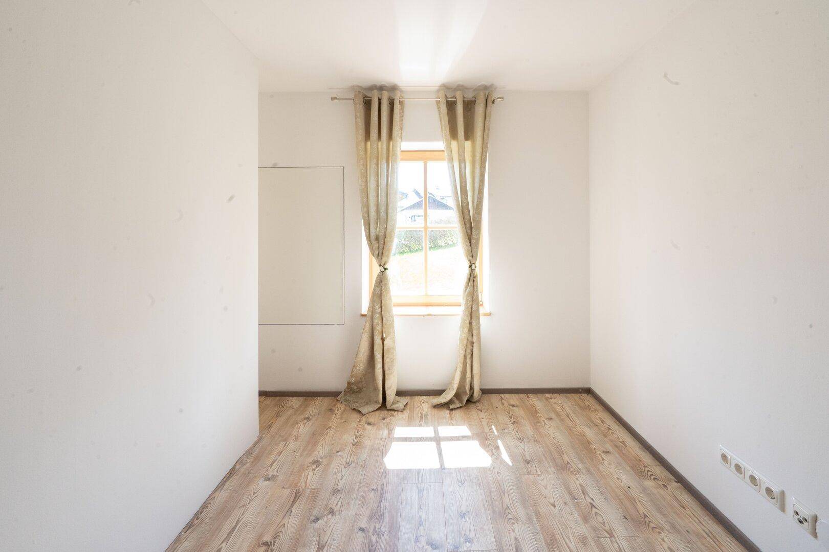 Leerer Raum mit Holzboden und Fenster, das viel Tageslicht hereinlässt.