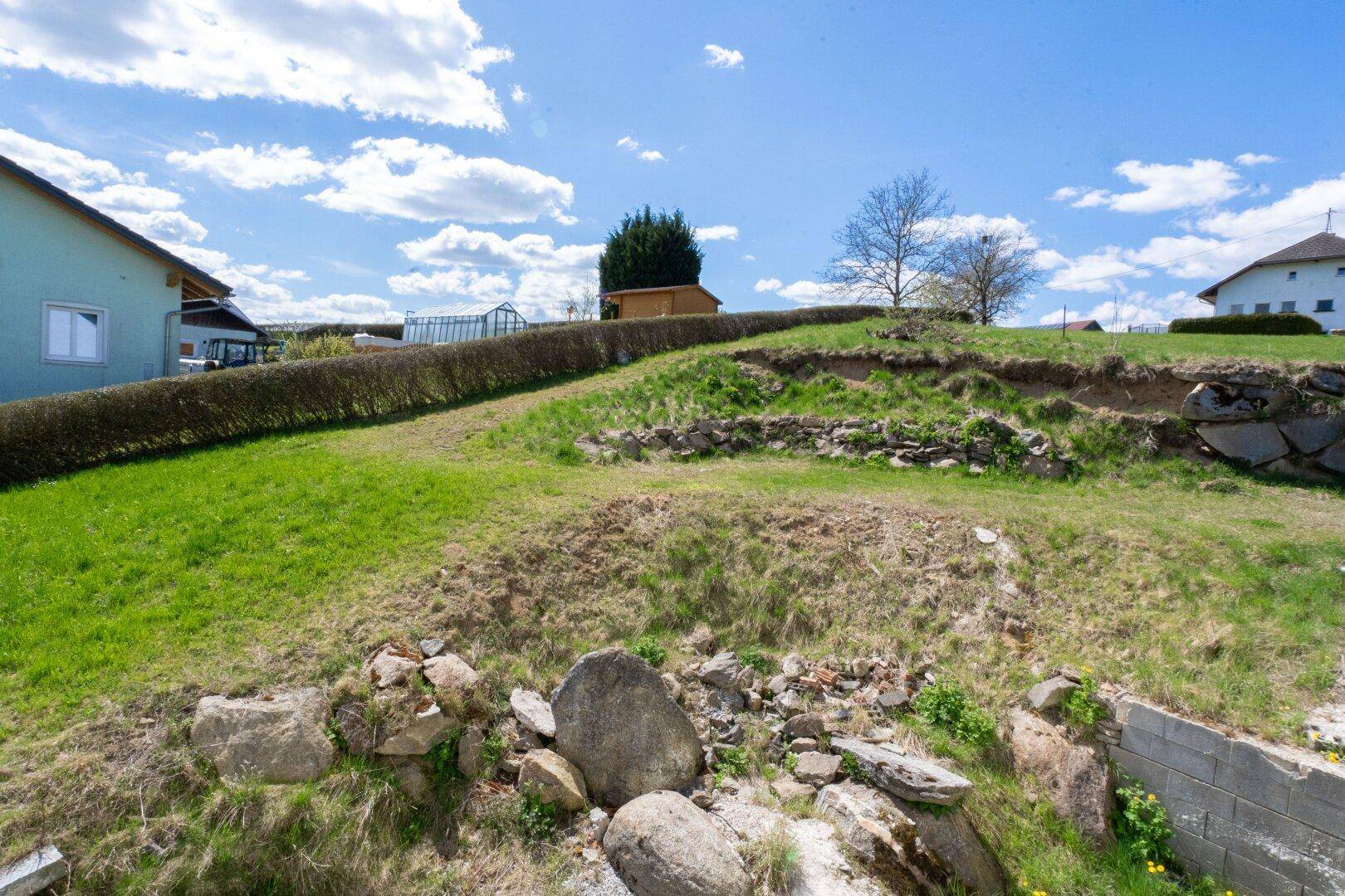 Grüne, leicht abfallende Gartenfläche mit Hecke und Felsen unter blauem Himmel.