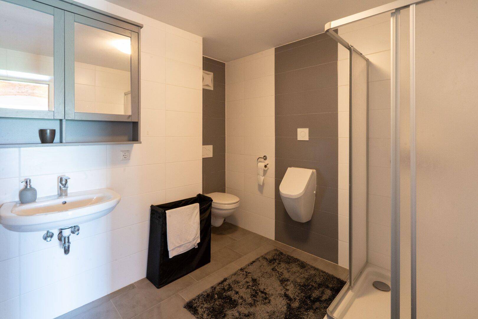 Modernes Badezimmer mit Dusche, Urinal und Waschbecken, ausgestattet mit hellen Fliesen.