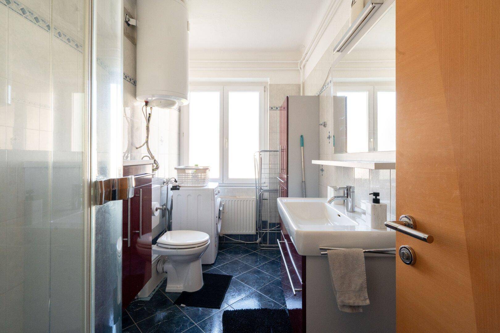 Badezimmer mit Dusche, WC, Waschbecken und Waschmaschine, hell durch großes Fenster.