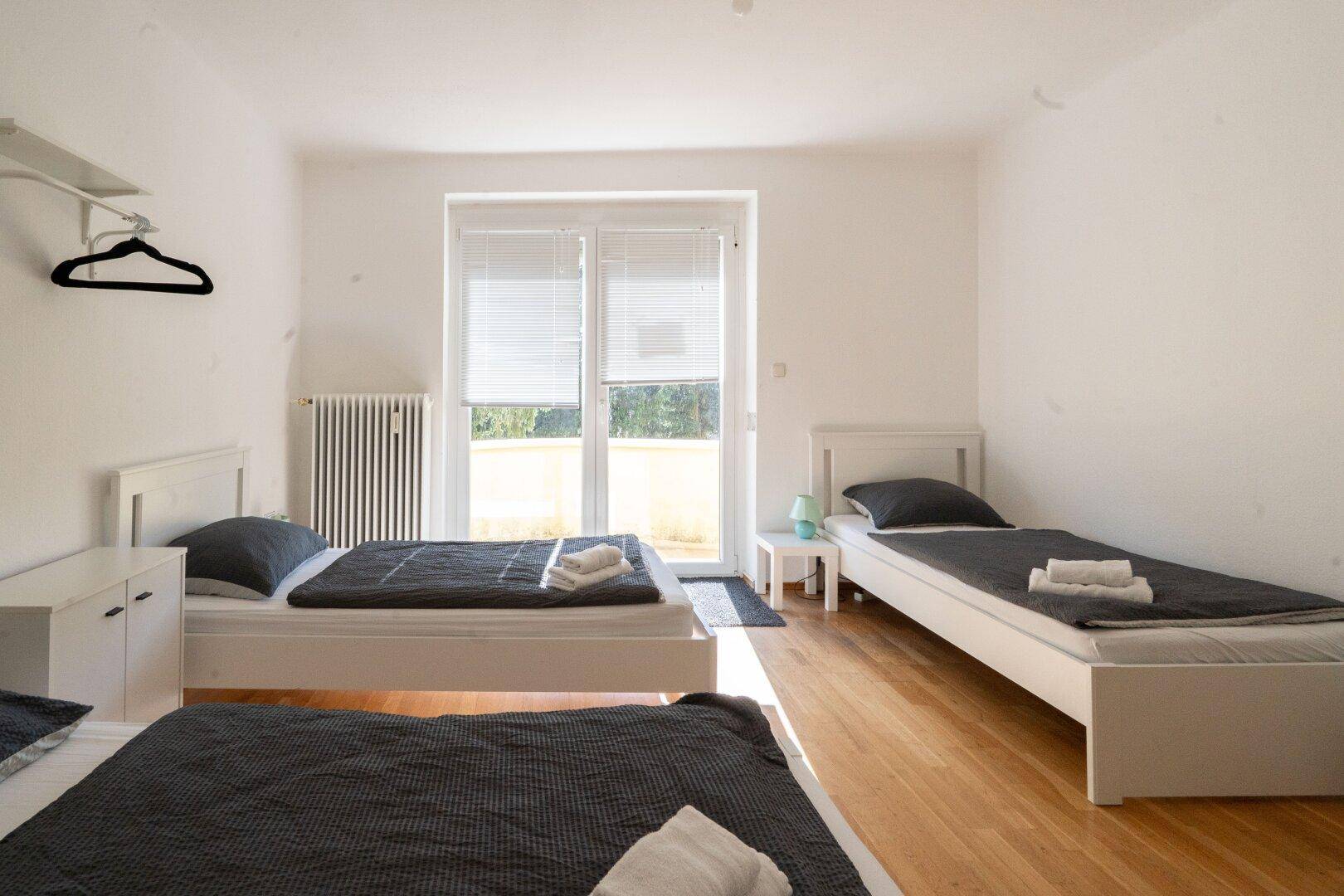 Helles Schlafzimmer mit drei Einzelbetten, Holzfußboden und Zugang zu einem Balkon.