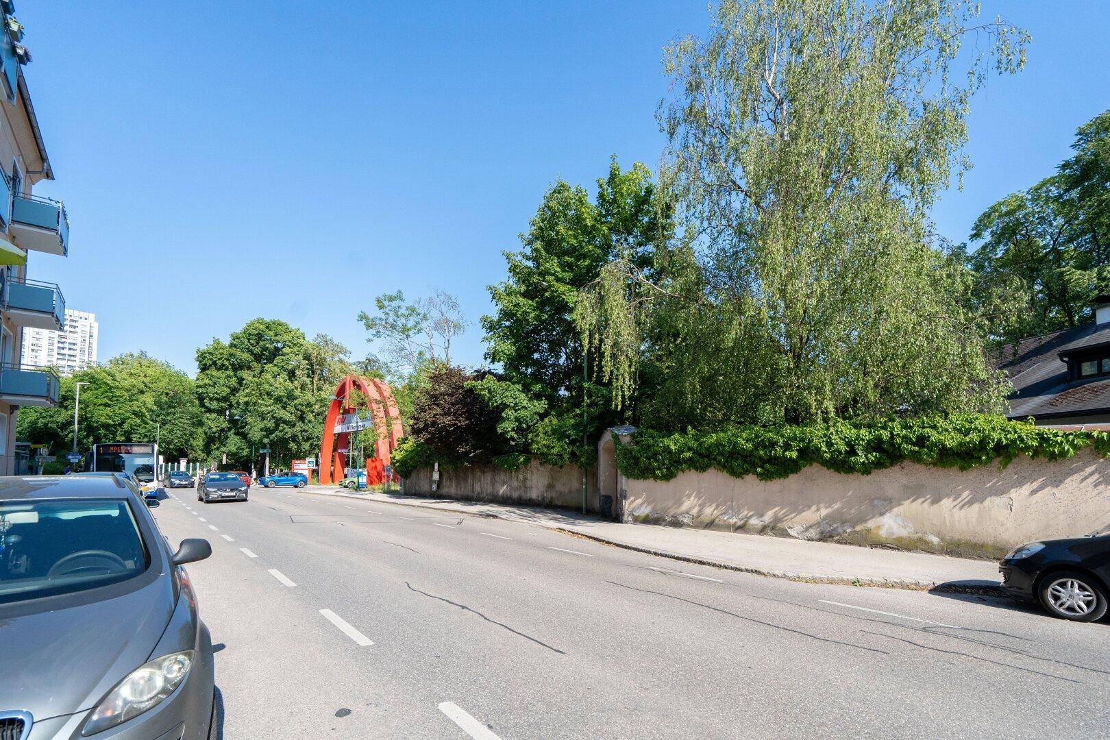 Straßenansicht mit parkenden Autos, Bäumen und einem markanten roten Torbogen im Hintergrund.